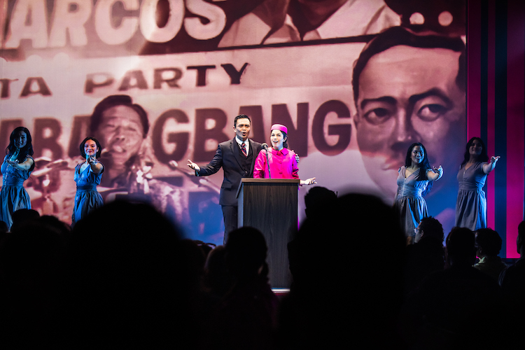 Jose Llana  and  Arielle Jacobs as Ferdinand and Imelda Marcos sing a victory speech in front of a huge black and white political poster of support for the dictator. In grey dresses a group of women reach out to the audience in solidarity. Imelda wears a shocking pink Chanel type suit with a Jackie O pillbox hat and Ferdinand looks dapper in black.